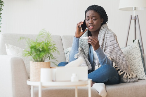 A young woman wrapped in a blanket on the couch looks at a thermometer.