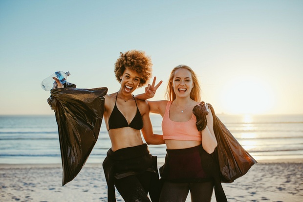 Two young women in swimming costumes and wetsuits smile on a beach while holding trash bags and rubbish